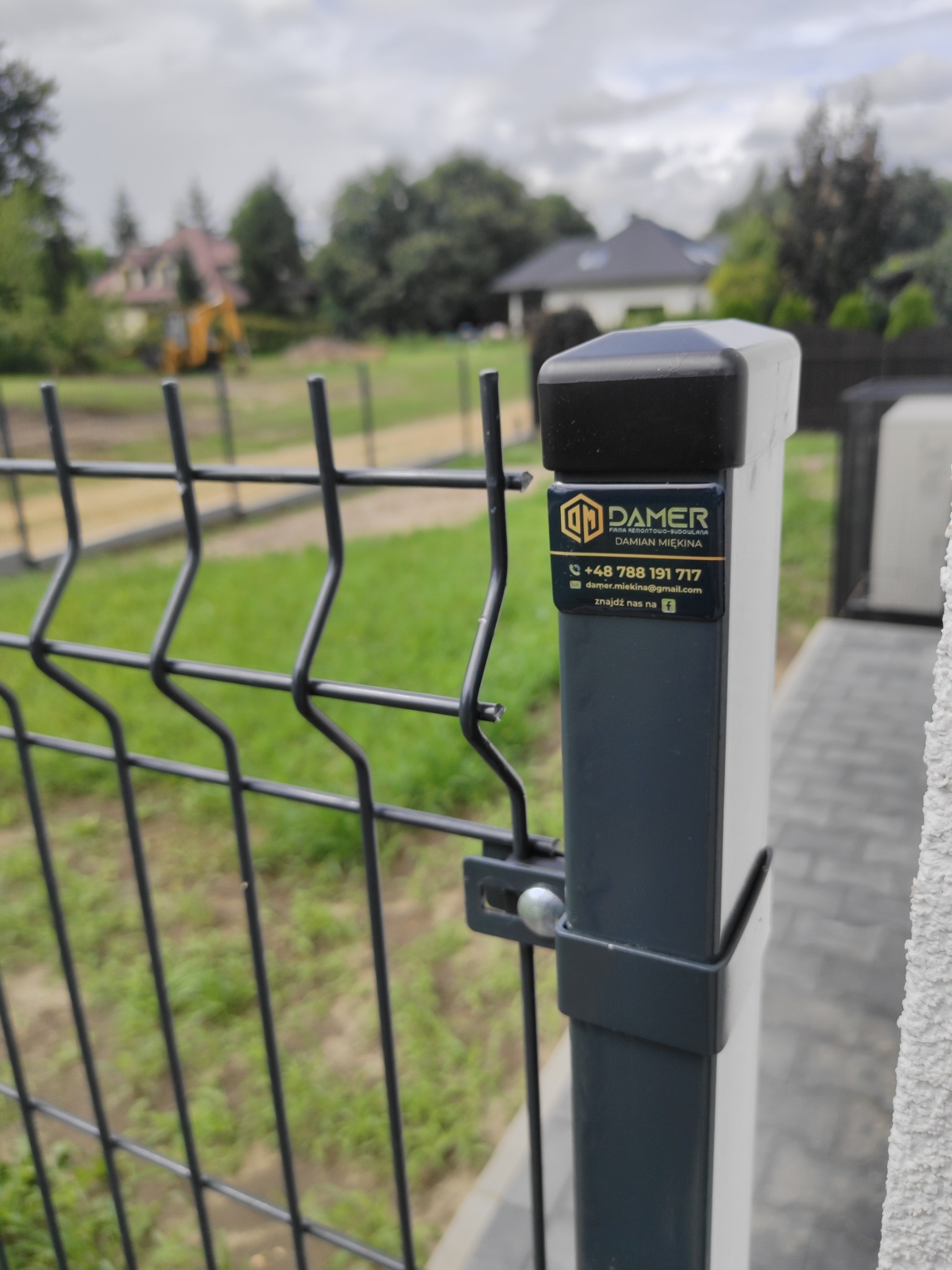 Fragment ogrodzenia panelowego z naklejoną wizytówką firmy remontowo-budowlanej DAMER, w tle zielony trawnik, dom i koparka.