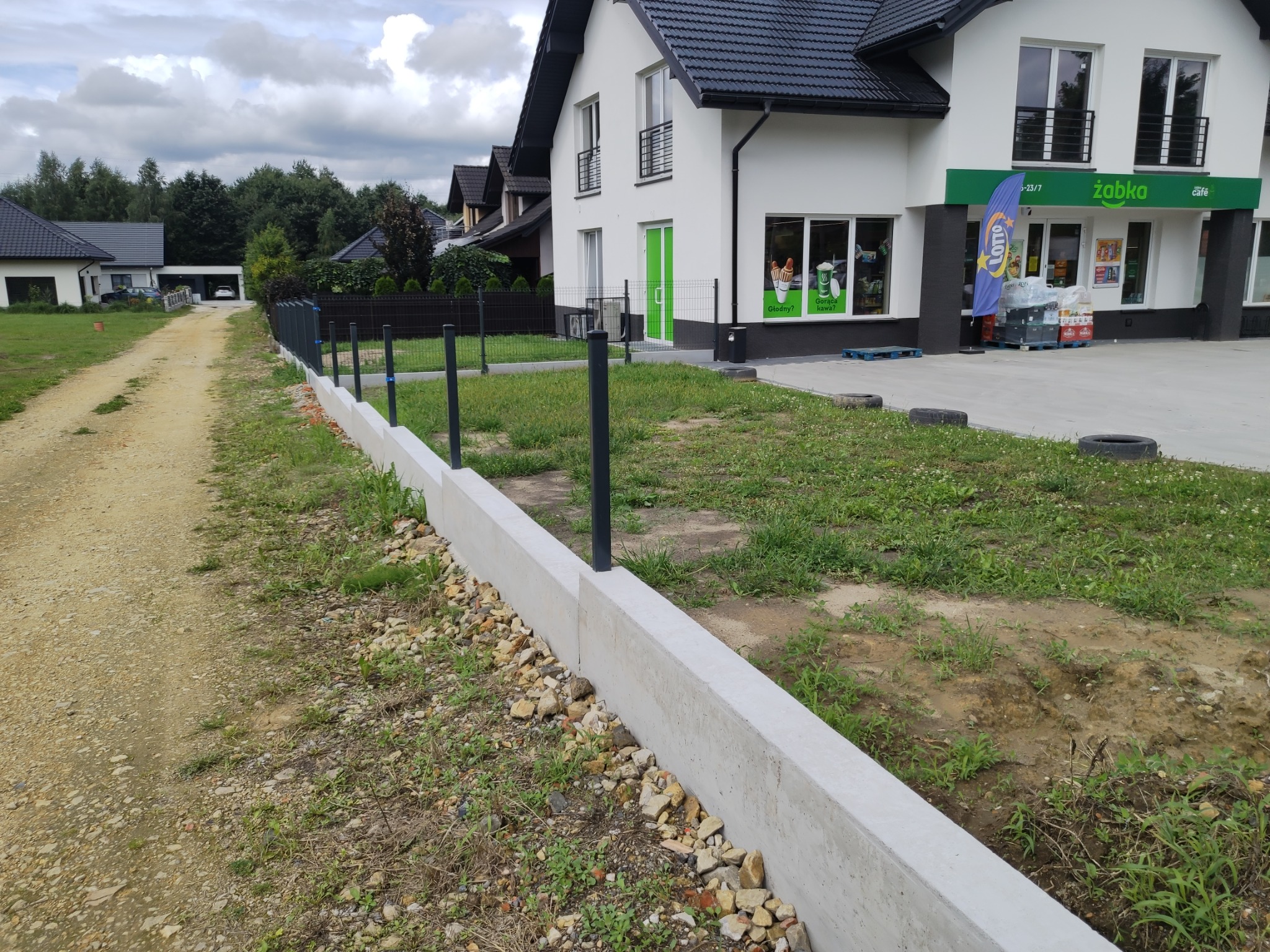 Świeżo wylany fundament pod ogrodzenie z zamontowanymi słupkami, widok na sklep Żabka i domy w tle.