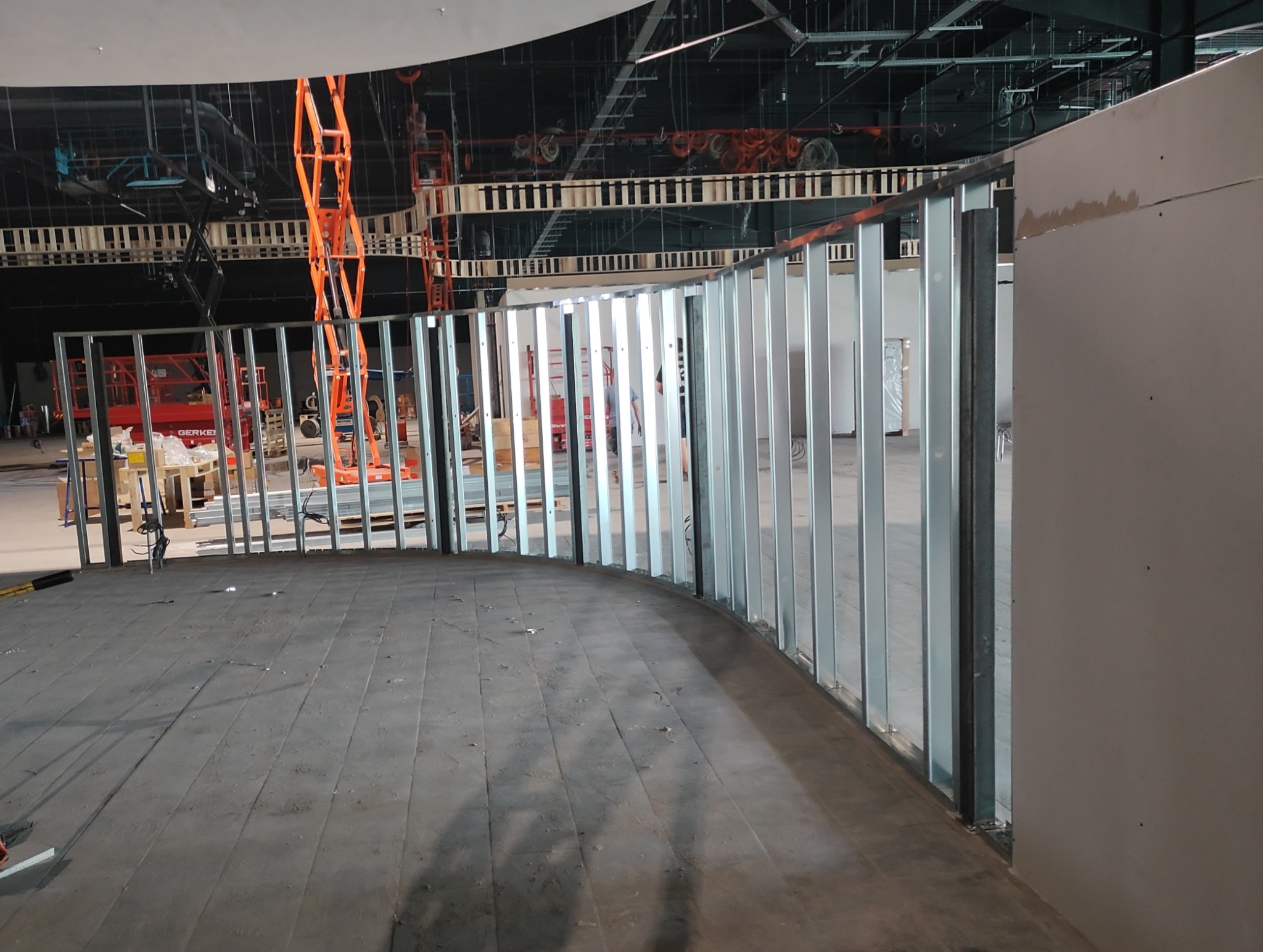 Konstrukcja ścianki działowej z profili stalowych w kształcie łuku, z częściowo zamontowaną płytą karton-gips, widoczne podnośniki nożycowe i rusztowania w tle przestrzeni komercyjnej w trakcie...