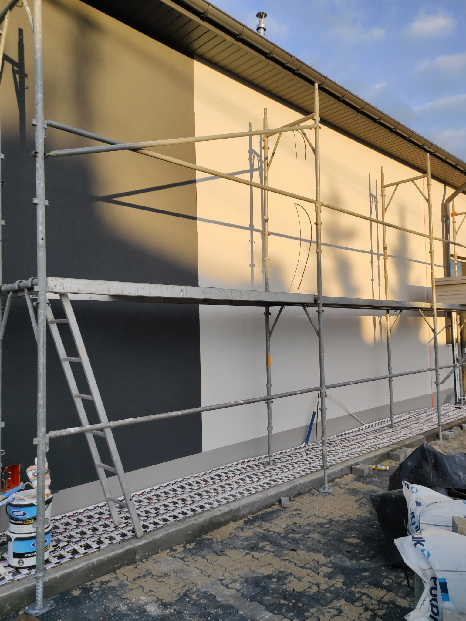 Elewacja budynku w trakcie malowania na dwa kolory - szary i biały, widoczne rusztowanie, puszki z farbą i zabezpieczona kostka brukowa.