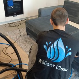 Brilliant Clean Paulina Jaworska - Pracownik w koszulce z logo firmy czyści ciemnoszarą kanapę za pomocą niebieskiego, profesjonalnego ekstraktora do tapicerki Santoemma Sabrina.