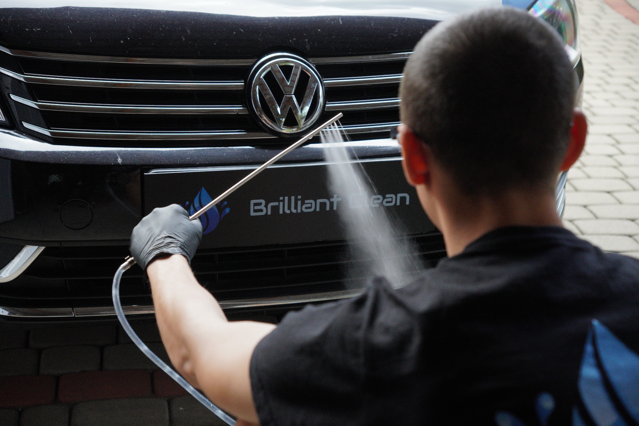Czyszczenie grilla samochodu Volkswagen za pomocą dyszy ciśnieniowej przez osobę w czarnych rękawicach, widoczny logotyp firmy Brilliant Clean.