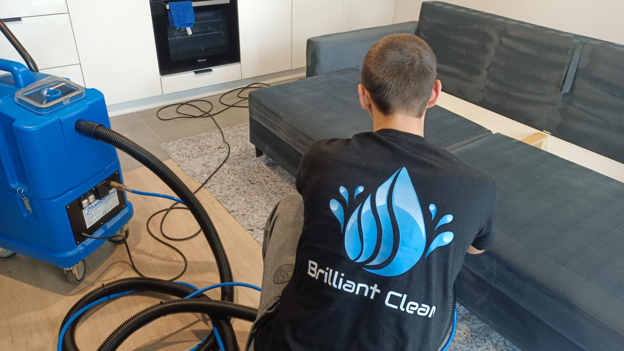 Pracownik w koszulce z logo firmy czyści ciemnoszarą kanapę za pomocą niebieskiego, profesjonalnego ekstraktora do tapicerki Santoemma Sabrina.