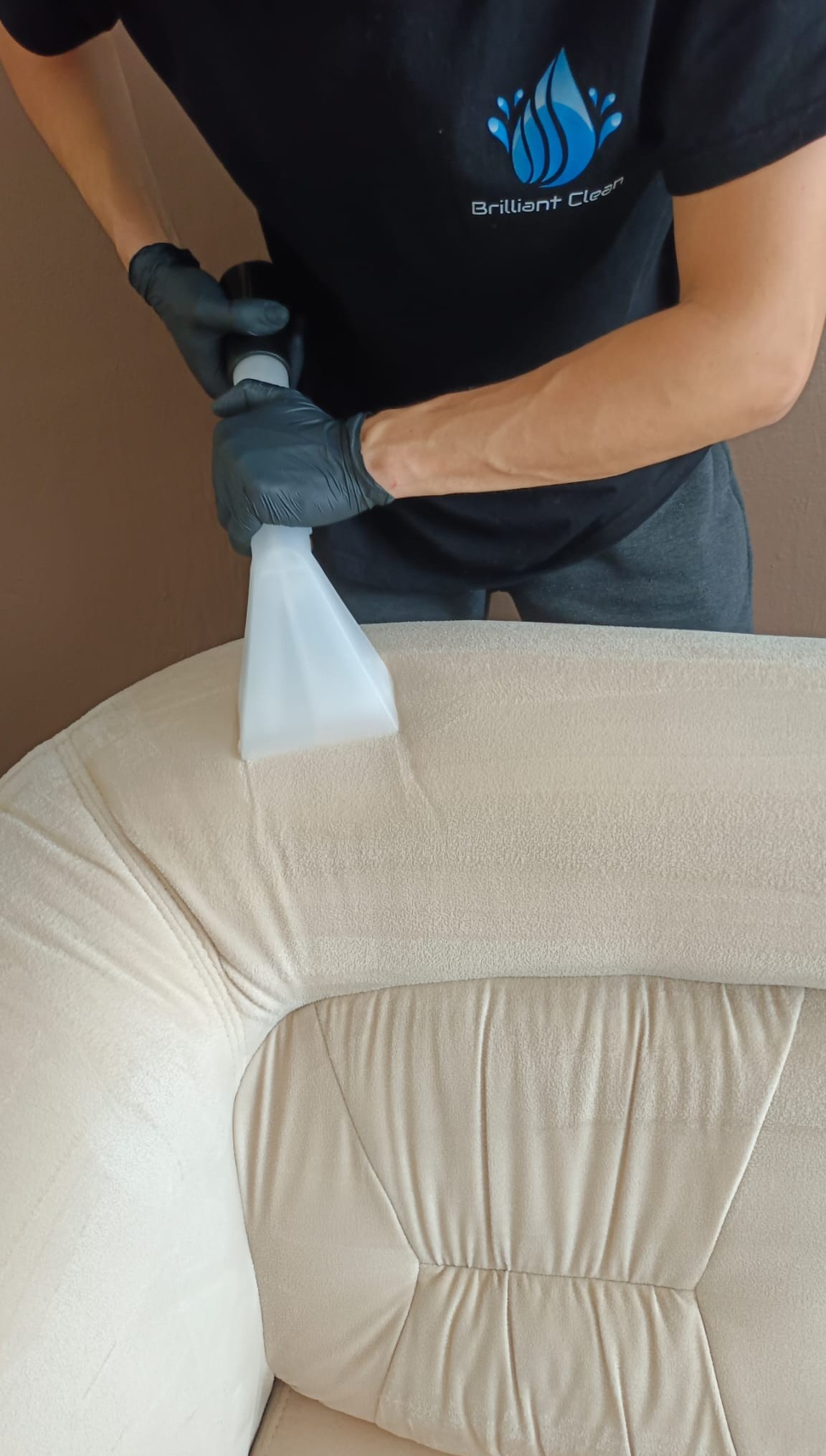 Pranie jasnej, pluszowej kanapy za pomocą ekstraktora; widoczna dysza i logo firmy Brilliant Clean na koszulce pracownika.