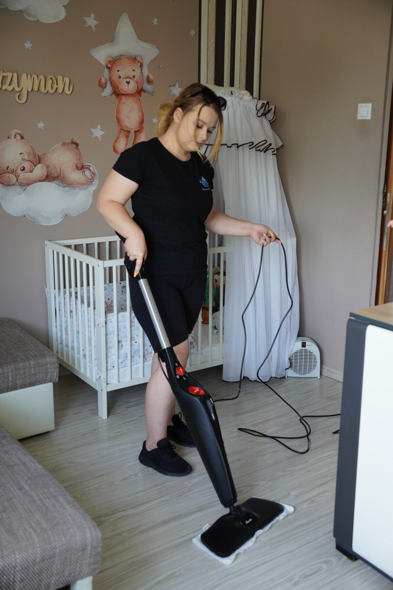 Kobieta w czarnym stroju czyści podłogę mopem parowym w pokoju dziecięcym z łóżeczkiem i dekoracjami ściennymi z misiami.