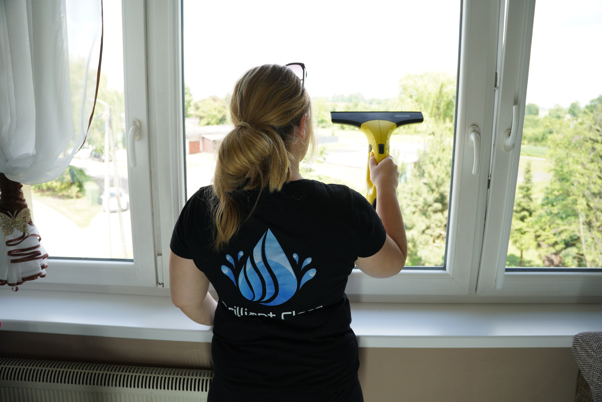 Kobieta w czarnej koszulce z logo firmy czyści okno za pomocą żółtej myjki Karcher, widok z tyłu.