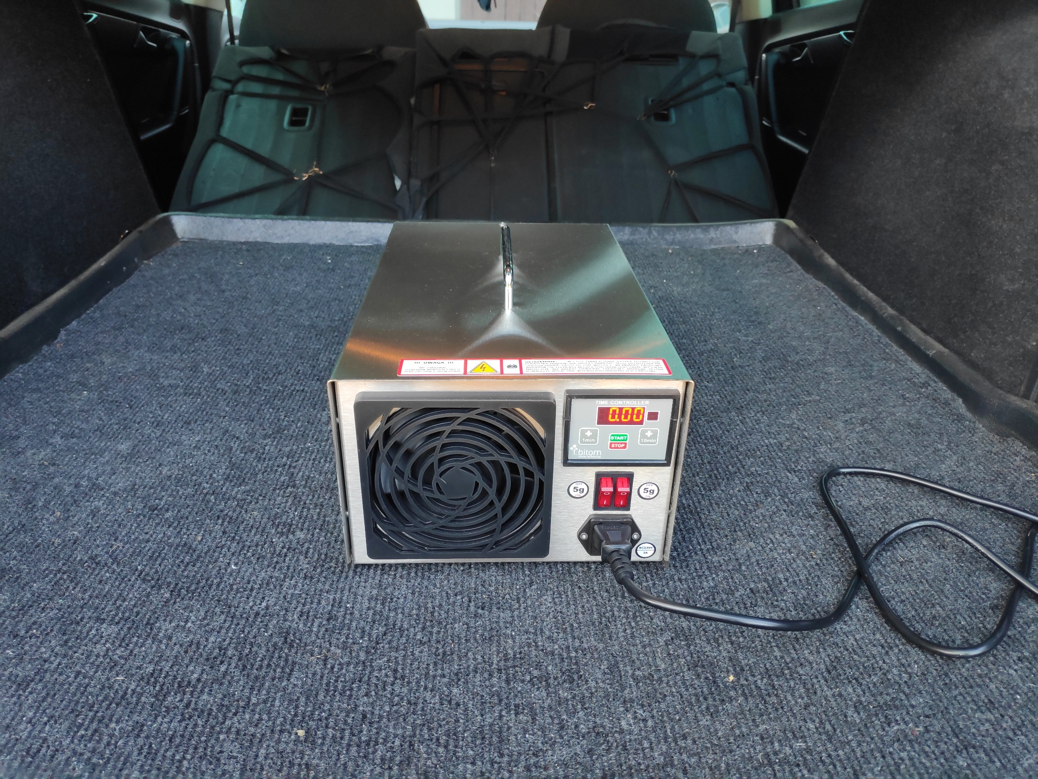 Ozonator w bagażniku samochodu, widoczny panel sterowania z wyświetlaczem, wentylator i kabel zasilający.
