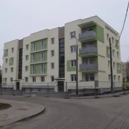 Nowoczesny budynek mieszkalny po remoncie elewacji, z zielonymi akcentami i balkonami, ogrodzony tymczasowym płotem budowlanym, widoczne sąsiednie budynki.