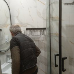 Radosław Olejnik - Mężczyzna w kurtce ogląda nową kabinę prysznicową z czarnymi profilami i armaturą w łazience wykończonej białymi płytkami imitującymi marmur.