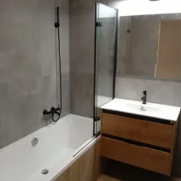 Nowoczesna łazienka z szarą płytką, białą wanną z parawanem nawannowym i drewnianą szafką pod umywalką z czarną armaturą.