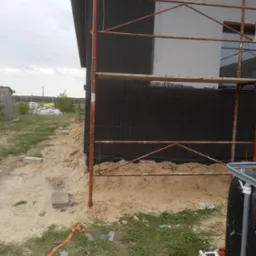Ściana budynku w trakcie prac elewacyjnych: fragment ocieplony czarnym styropianem, fragment z białym tynkiem, widoczne metalowe rusztowanie, piasek i gruz wokół budynku.