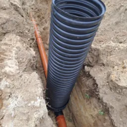 Wykop z ułożonymi rurami drenarskimi: czarna karbowana pionowa rura oraz pomarańczowa gładka pozioma rura. Widoczne korzenie i źdźbła trawy na ścianach wykopu.