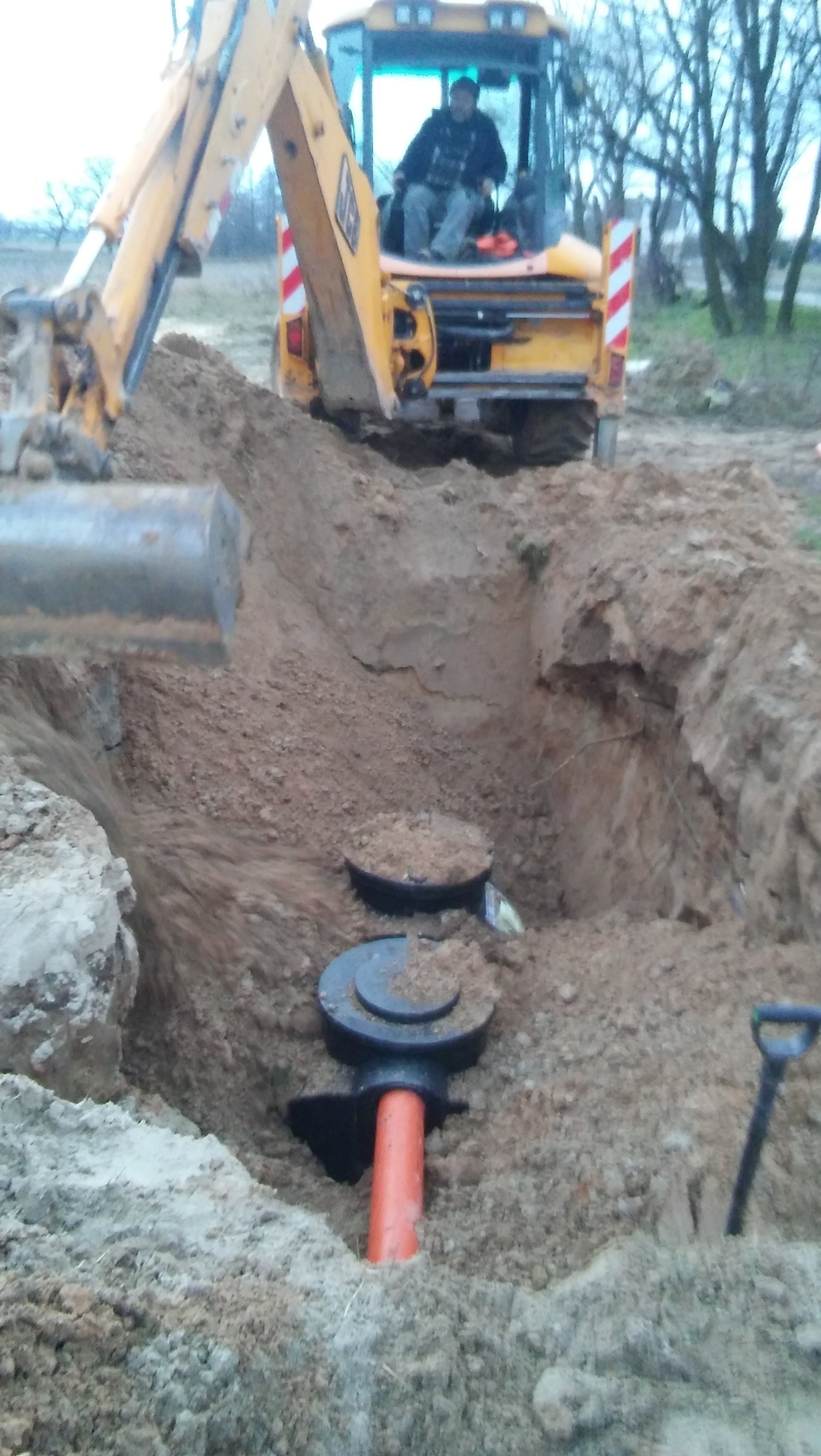 Żółta koparka przy wykopie z czarnymi elementami instalacji i pomarańczową rurą. W tle operator maszyny i drzewa.