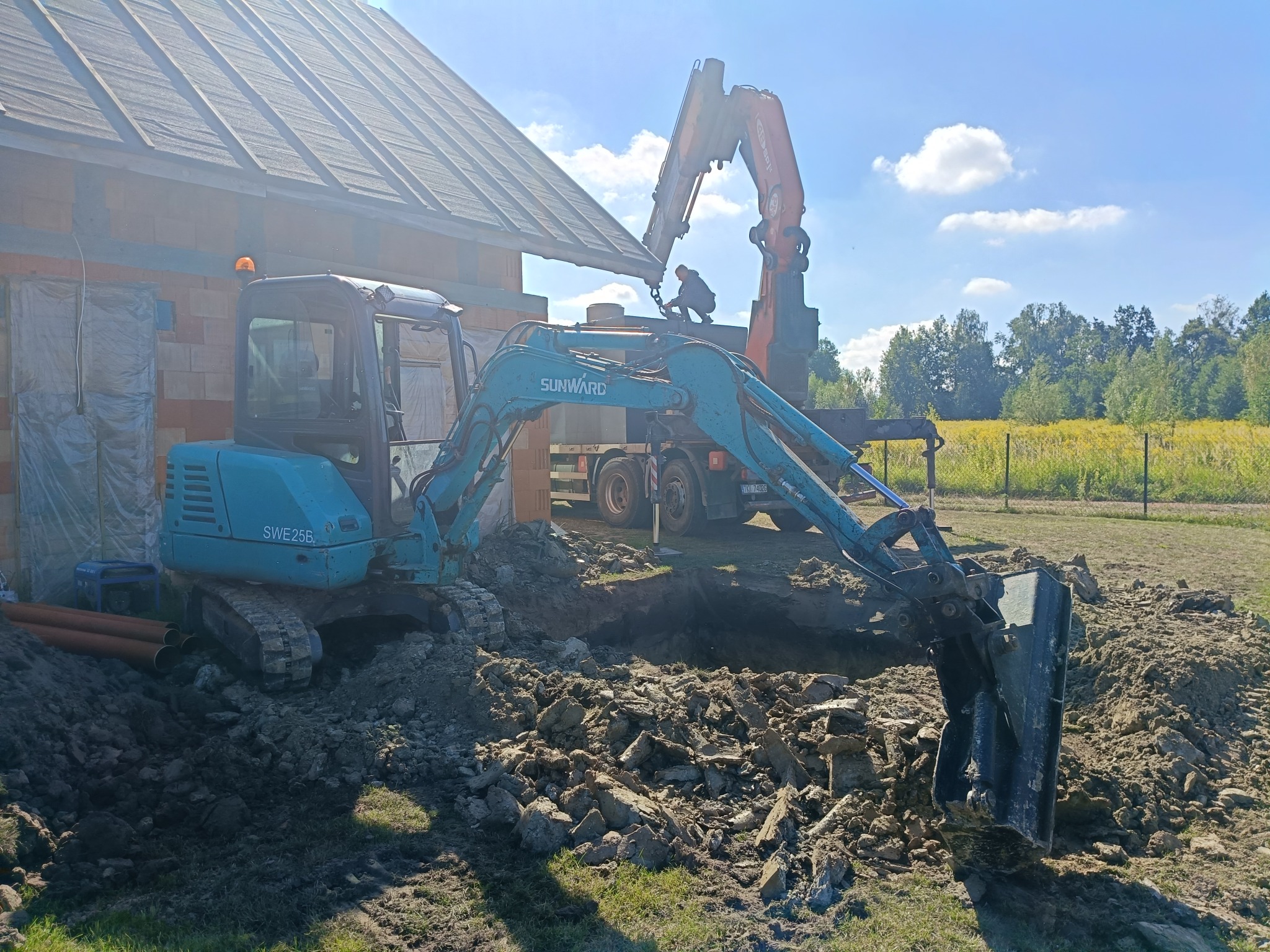 Niebieska mini koparka SUNWARD SWE25B wykonuje wykop. W tle budynek w trakcie budowy i ciężarówka z dźwigiem. Słoneczny dzień.