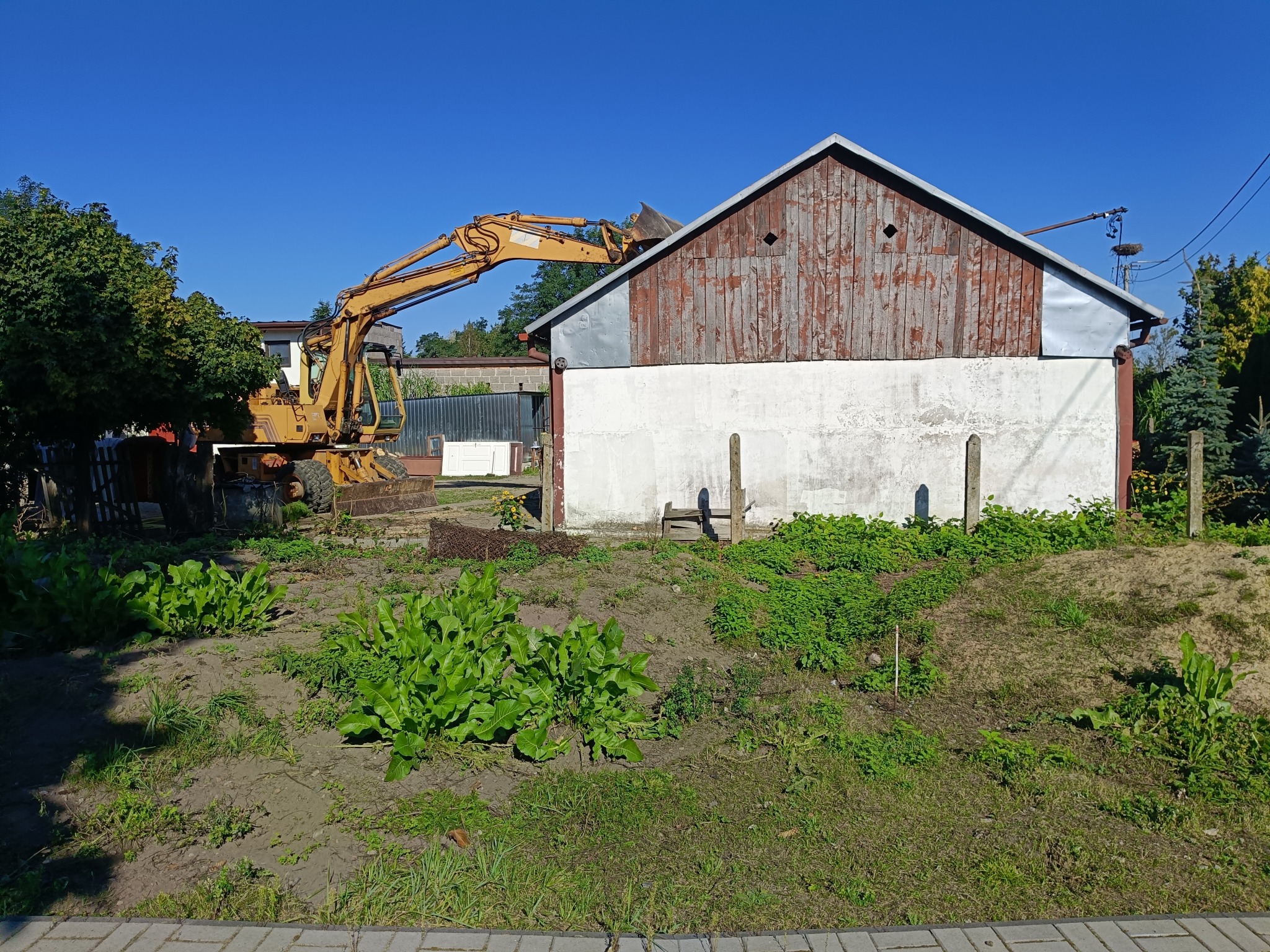 Żółta koparka mechanicznie rozbiera drewniany budynek z białą ścianą, otoczony zielenią w słoneczny dzień. Widoczne ślady wcześniejszych prac.