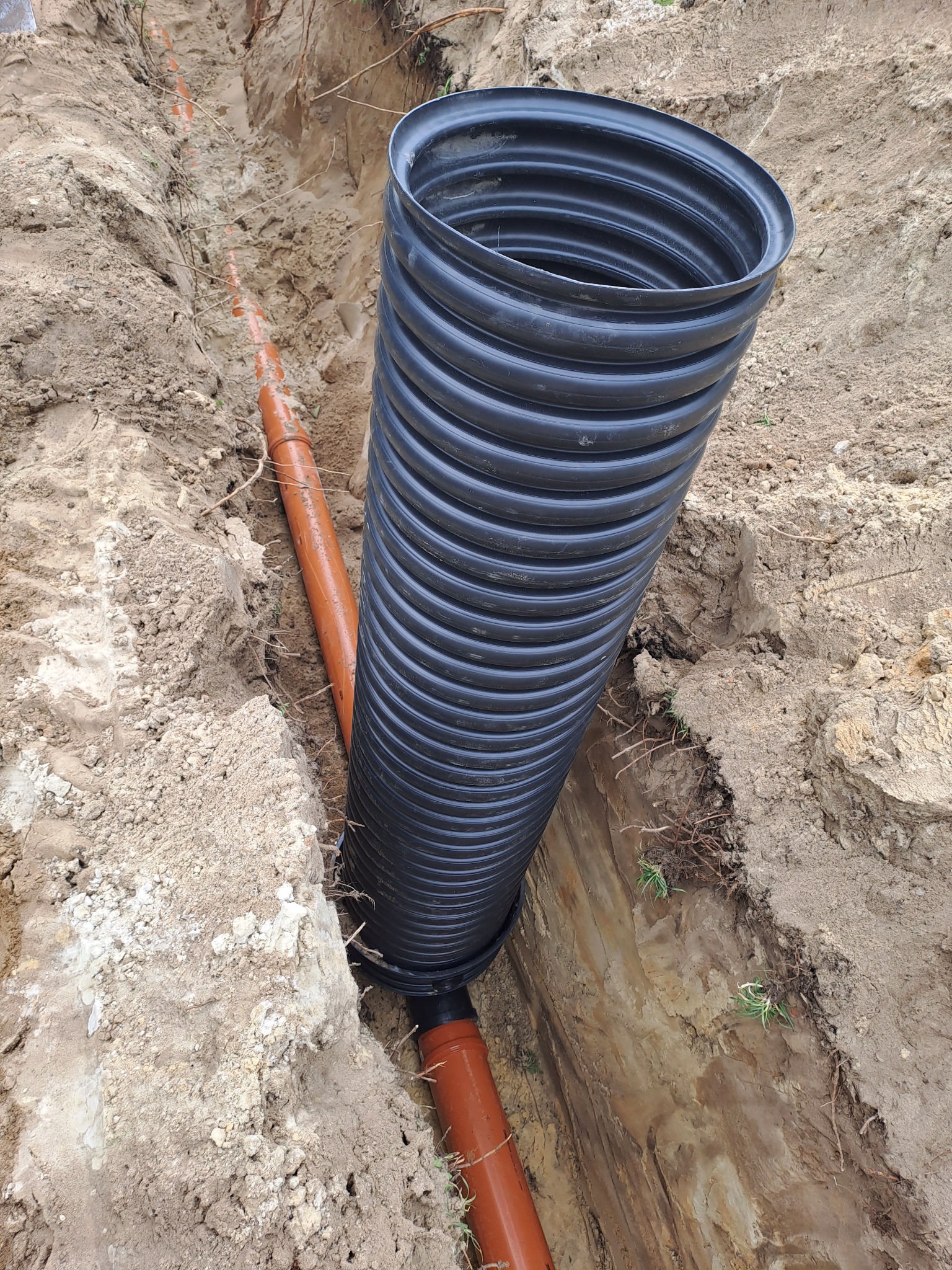 Wykop z ułożonymi rurami drenarskimi: czarna karbowana pionowa rura oraz pomarańczowa gładka pozioma rura. Widoczne korzenie i źdźbła trawy na ścianach wykopu.