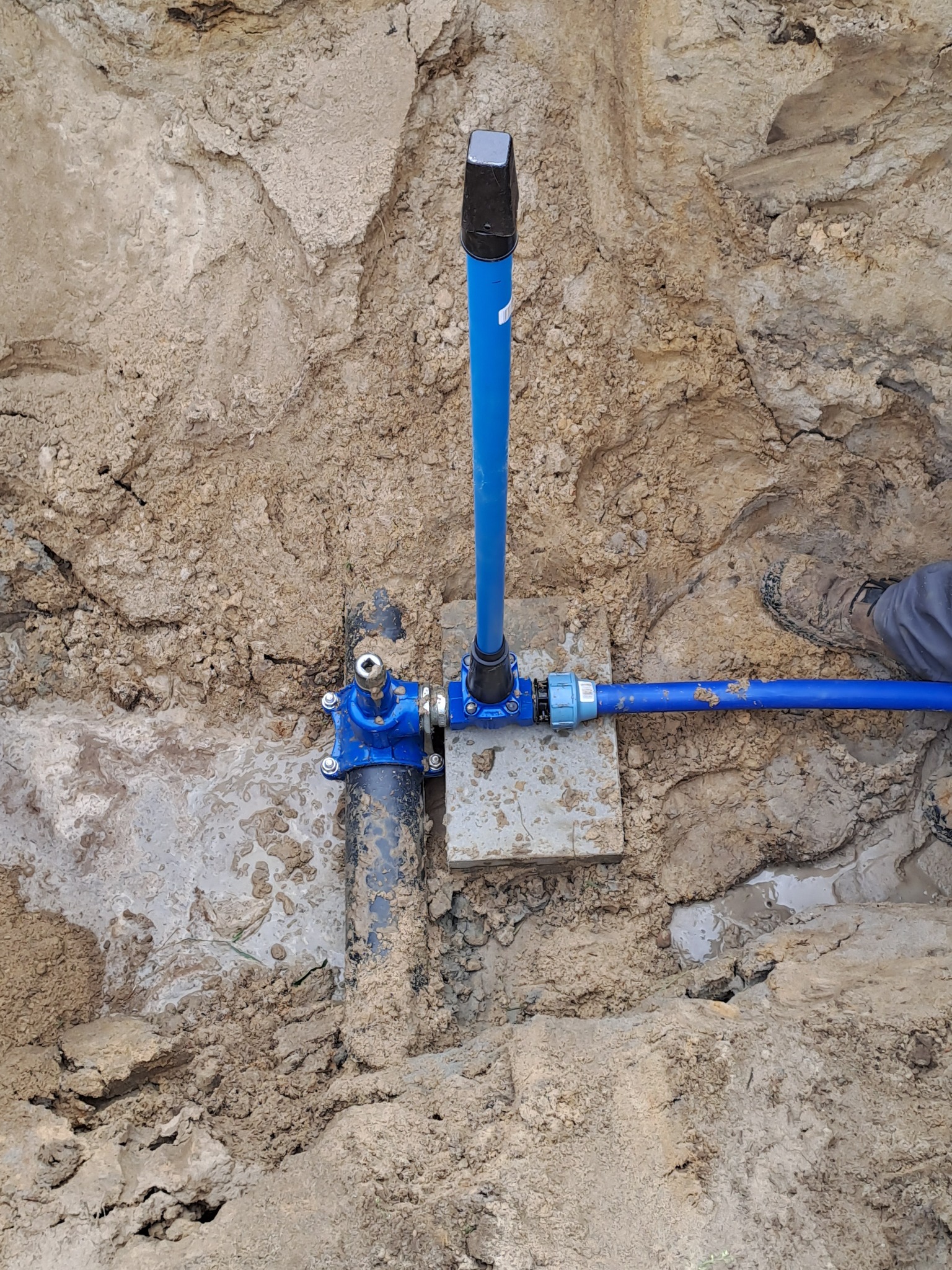 Niebieska rura instalacyjna wystająca z wykopu w ziemi, połączona z czarną rurą za pomocą niebieskiego zaworu, na ziemi leży betonowa płytka, widoczny fragment buta robotnika.
