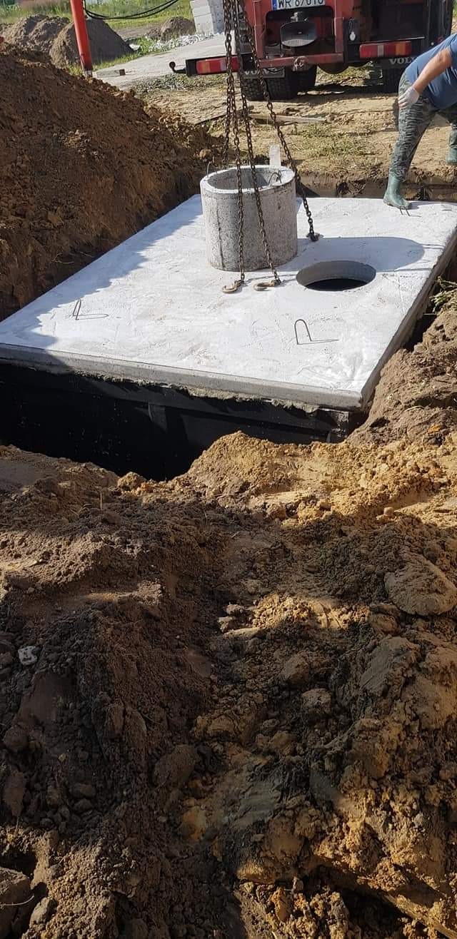 Montaż betonowego zbiornika: płyta z otworem opuszczana do wykopu za pomocą łańcuchów, widoczny fragment ciężarówki i pracownik w gumowych butach.