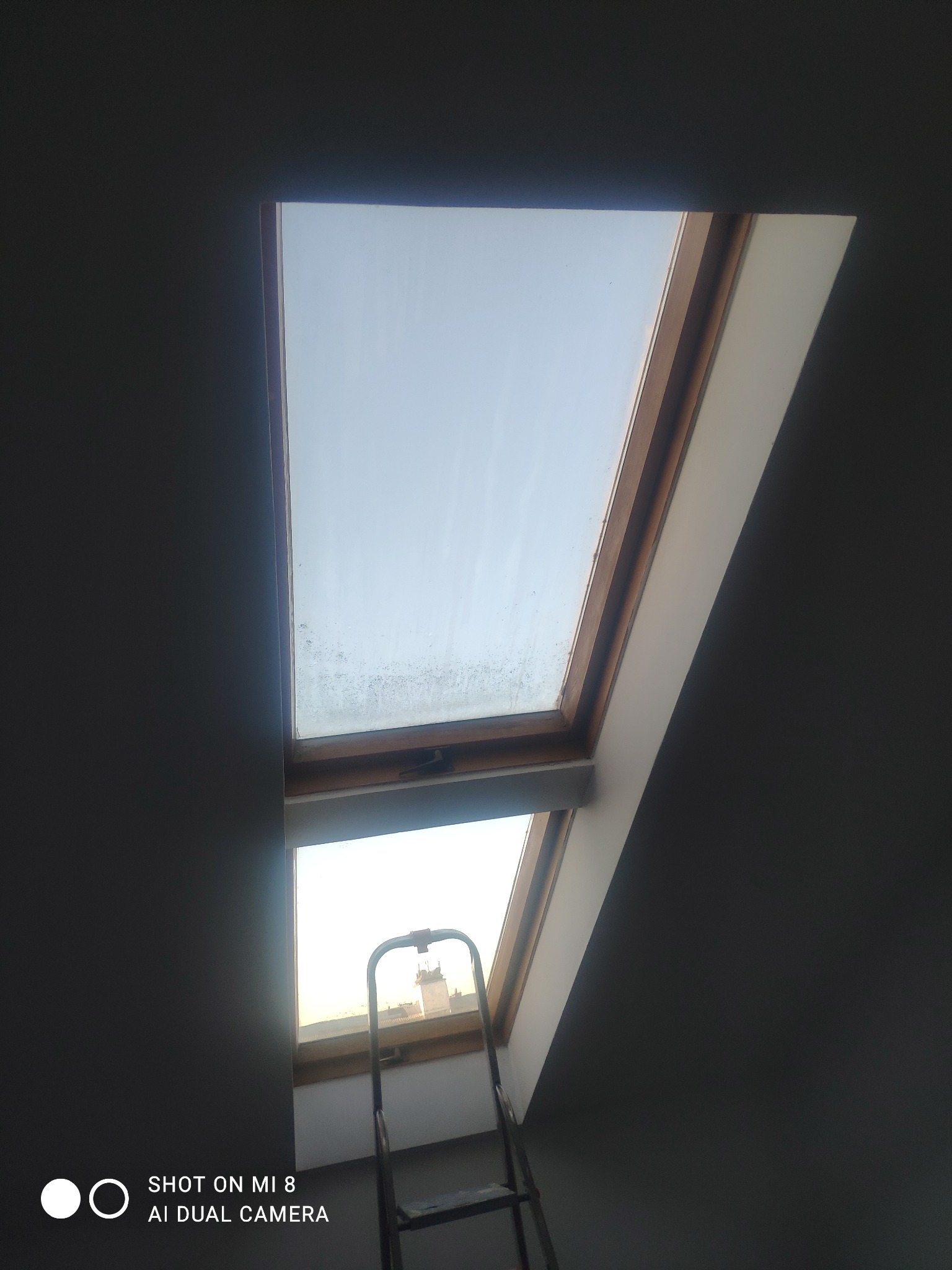 Poddasze z dwoma oknami dachowymi, dolne okno z drabinką opartą o parapet i widokiem na budynek w oddali, górne okno z drewnianą ramą i zabrudzeniami na szybie.