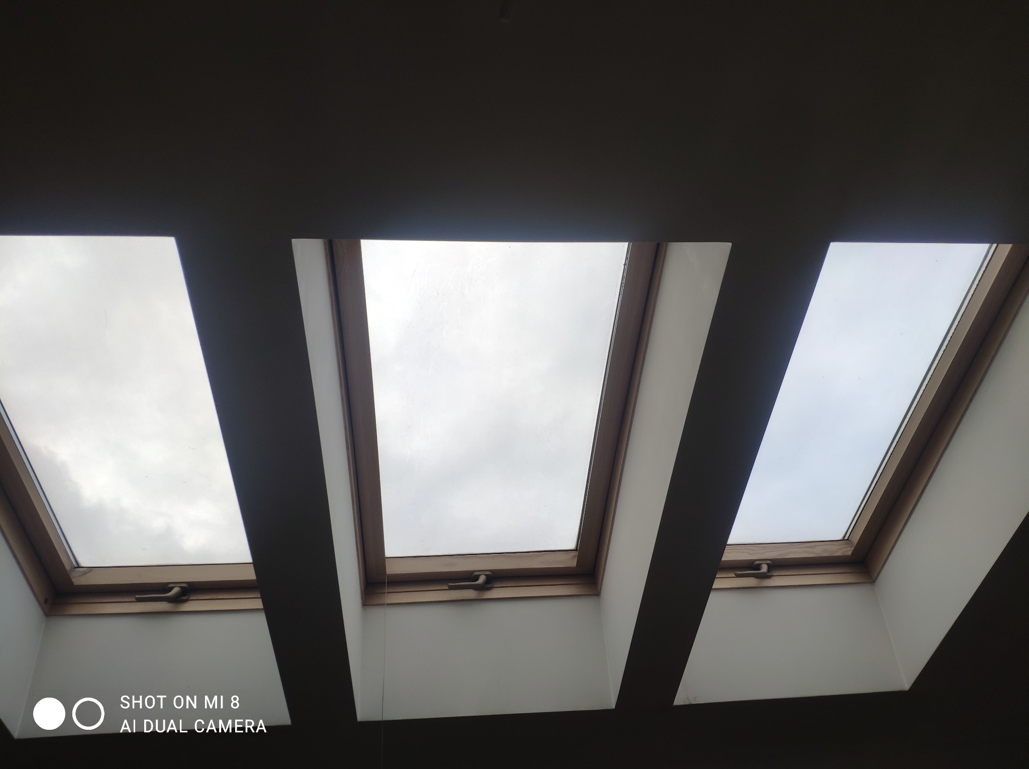 Trzy okna dachowe z drewnianymi ramami, widok z dołu na pochmurne niebo.