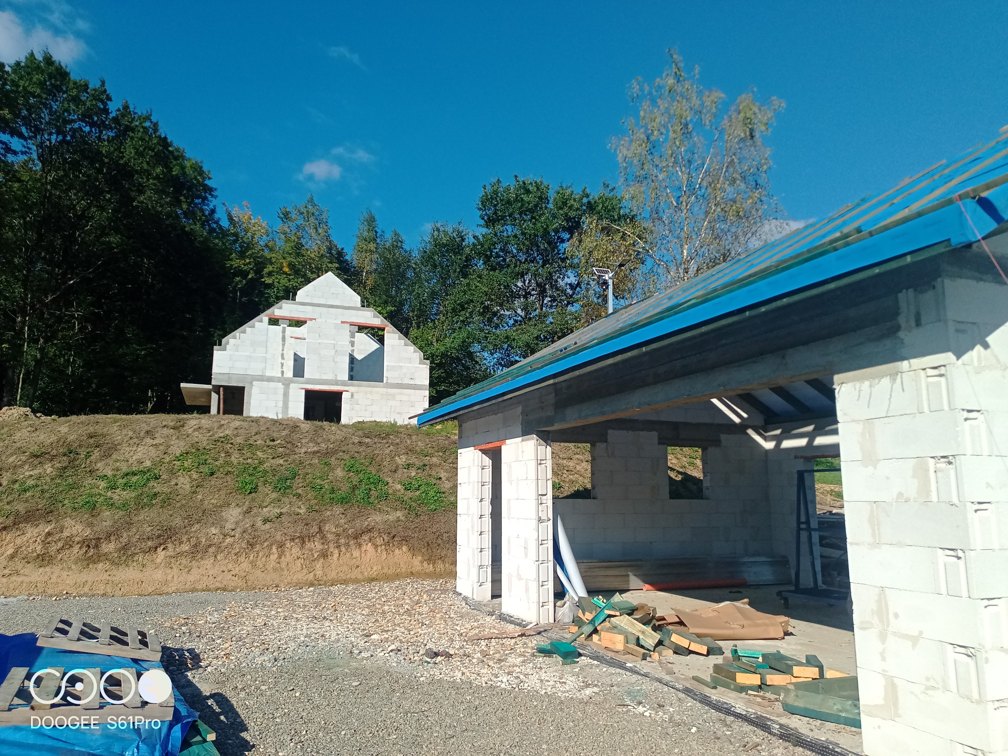 Budowa dwóch domów murowanych z bloczków betonowych na działce z widokiem na las, jeden dom w stanie surowym otwartym, drugi z dachem pokrytym niebieską folią, widoczne materiały budowlane na ziemi.