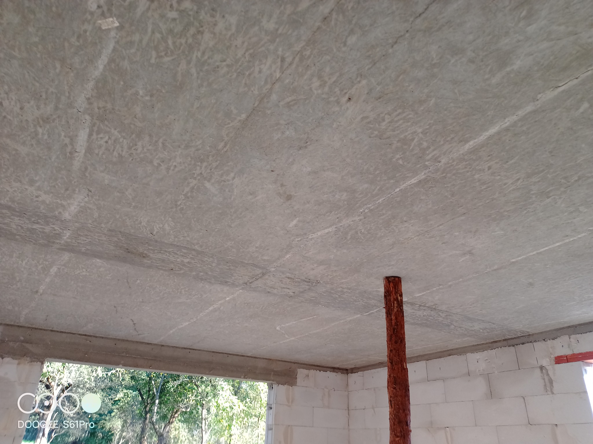 Surowy betonowy strop w trakcie budowy domu murowanego, wsparty drewnianą podporą, widok na zewnątrz przez otwór okienny.