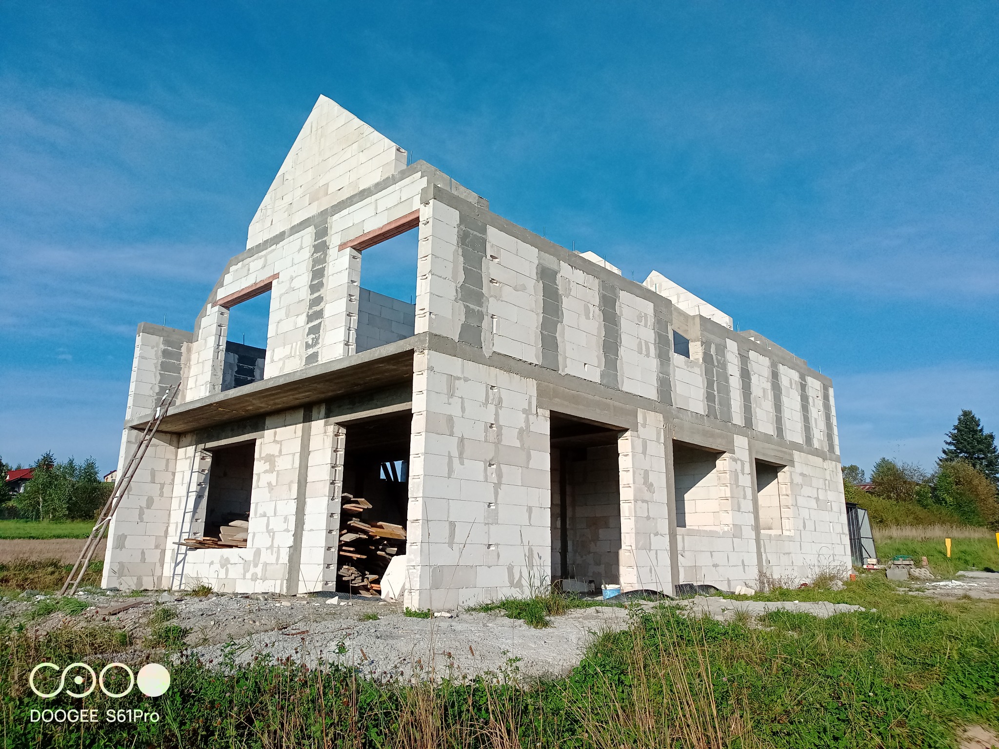 Niedokończony dwukondygnacyjny dom z białych bloczków, widoczne otwory okienne i drzwiowe, drewniana drabina oparta o ścianę, stos desek wewnątrz budynku, błękitne niebo w tle.