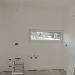 Wnętrze w trakcie prac przygotowawczych do wykończenia: ściany po gruntowaniu, widoczne puszki elektryczne, instalacja wodna, drabina narzędziowa i okno z widokiem na ogród.