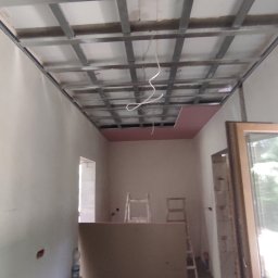 Serwis Remont - Montaż sufitu podwieszanego: metalowa konstrukcja kratowa zamocowana do stropu, częściowo obłożona płytą karton-gips w kolorze różowym, widoczne kable instalacji elektrycznej, w tle drabiny malarskie.