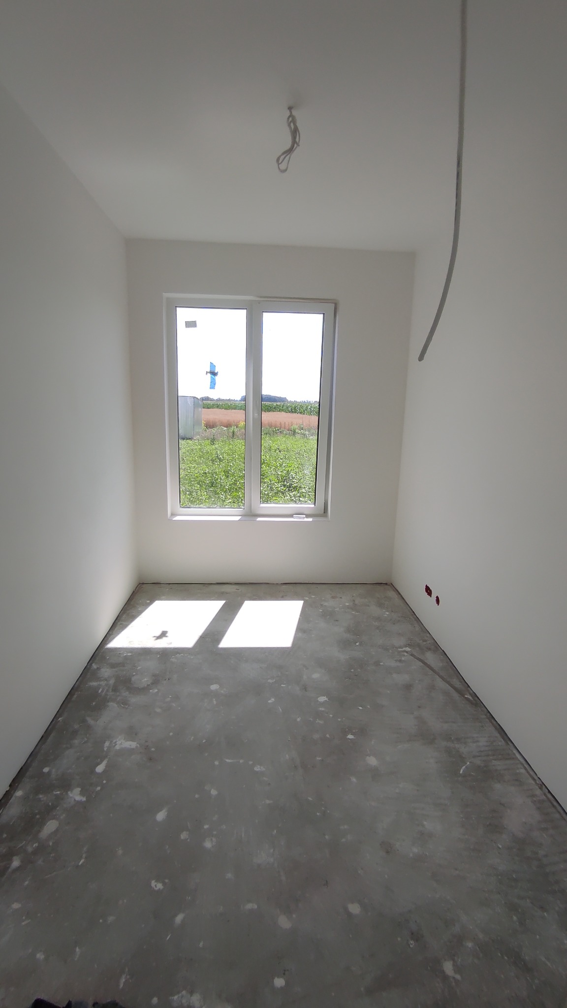 Pusty pokój z białymi ścianami, surowym betonowym podłożem i oknem wychodzącym na zielone pole. Widoczne wystające kable i niedokończone gniazdka elektryczne.