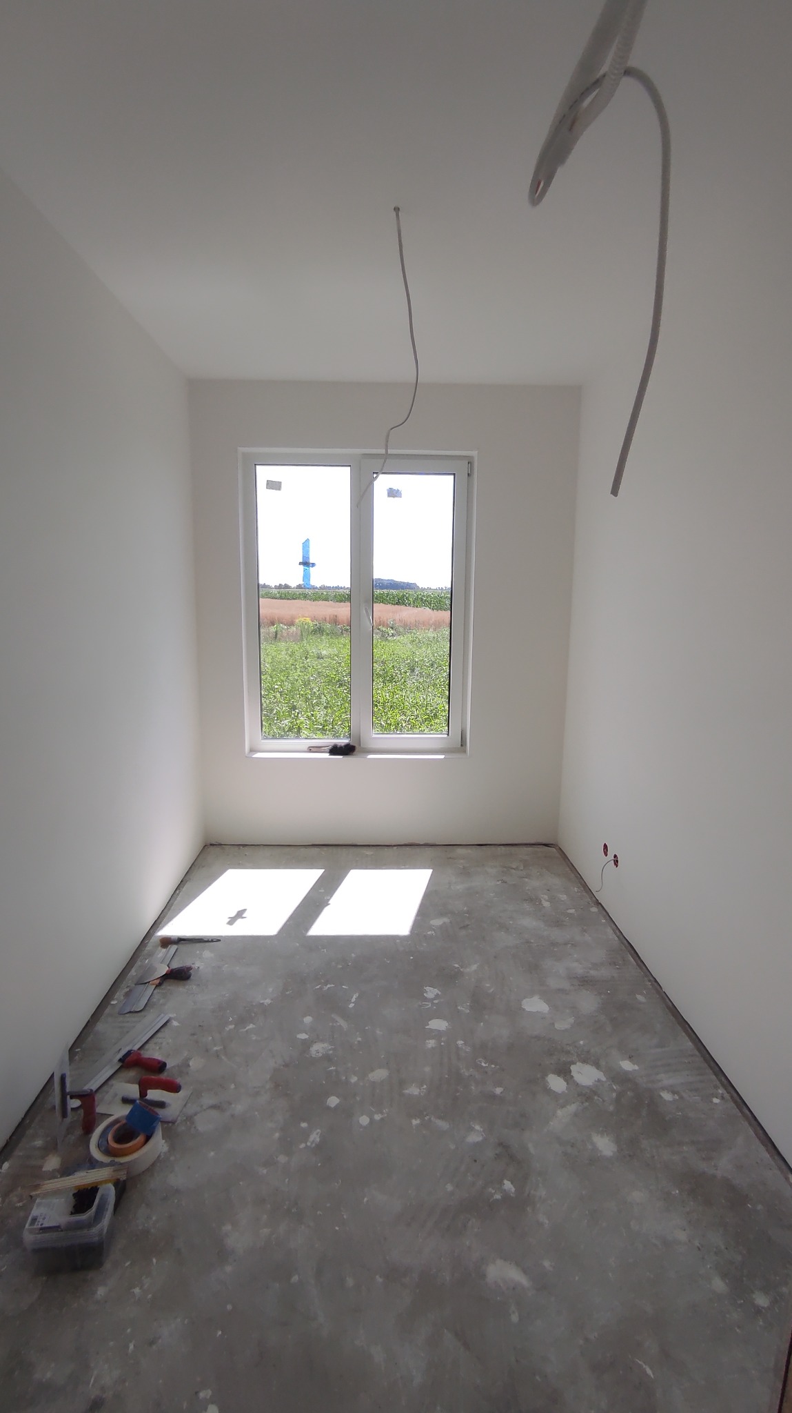 Pusty pokój po szpachlowaniu ścian, z widokiem na pole przez okno, narzędzia do szpachlowania leżące na podłodze, wystające przewody elektryczne z sufitu i gniazdka w ścianie.
