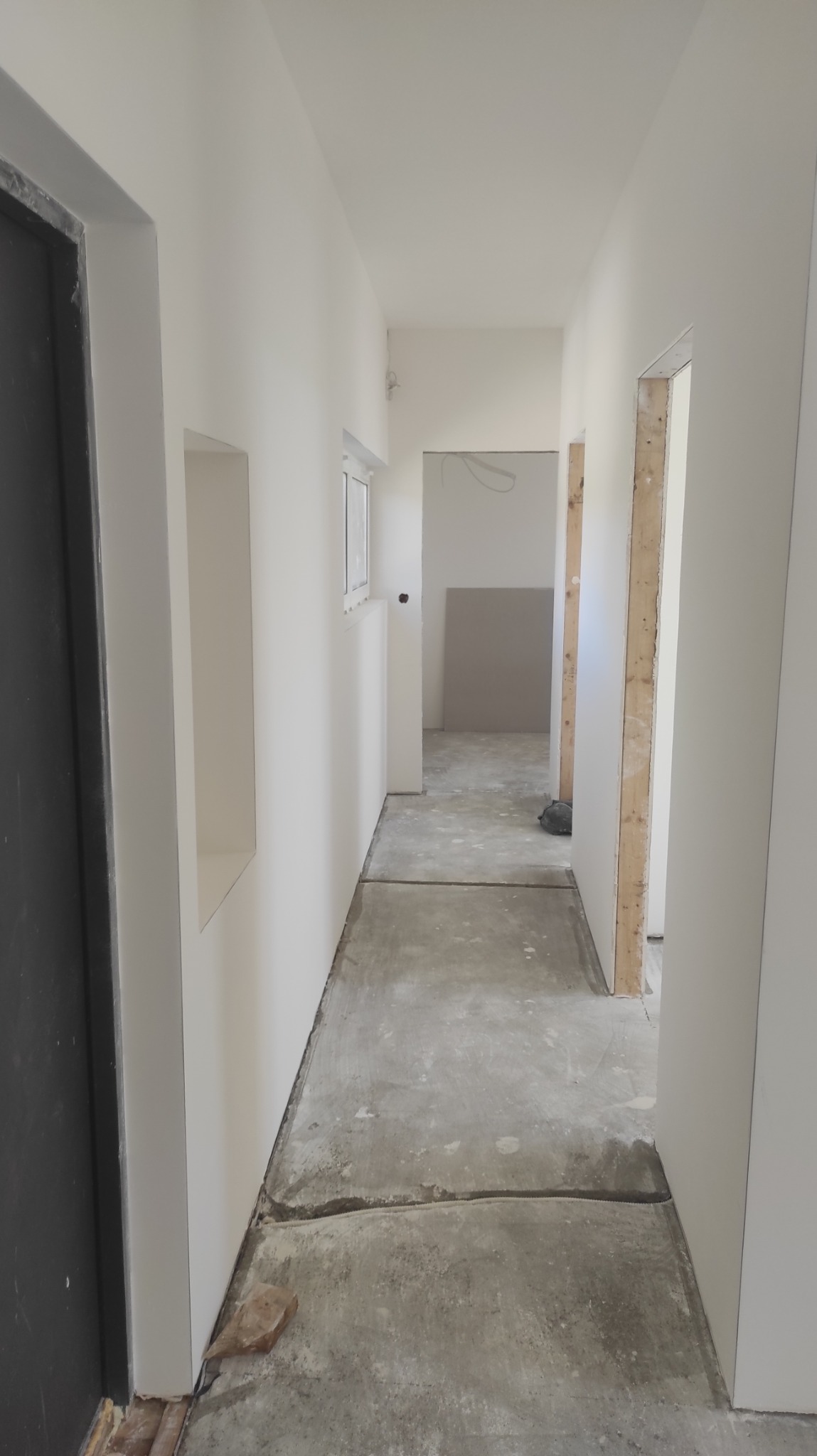 Korytarz w trakcie remontu z przygotowanymi ścianami do malowania, widoczna wnęka w ścianie, surowa podłoga i fragmenty konstrukcji drzwi.
