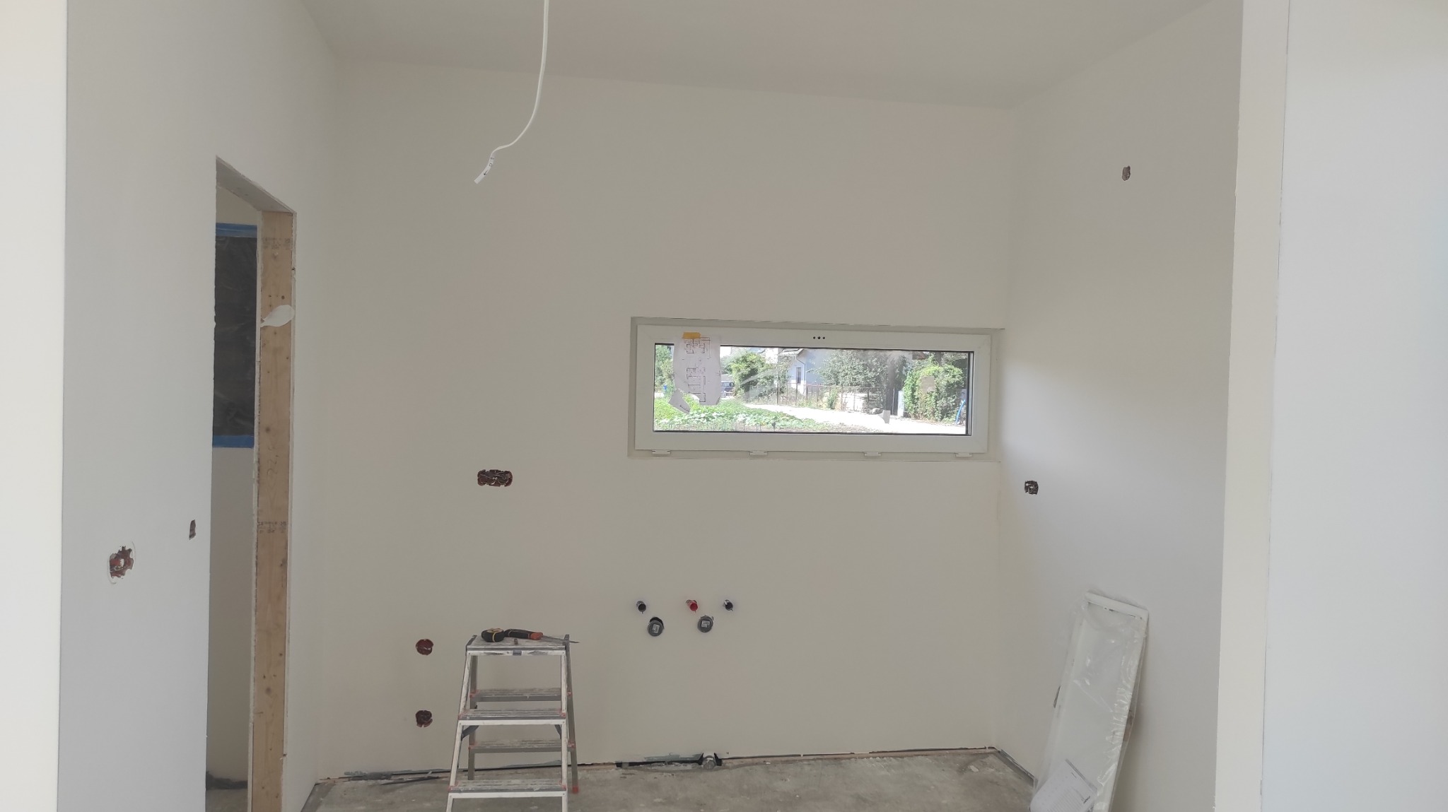 Wnętrze w trakcie prac przygotowawczych do wykończenia: ściany po gruntowaniu, widoczne puszki elektryczne, instalacja wodna, drabina narzędziowa i okno z widokiem na ogród.