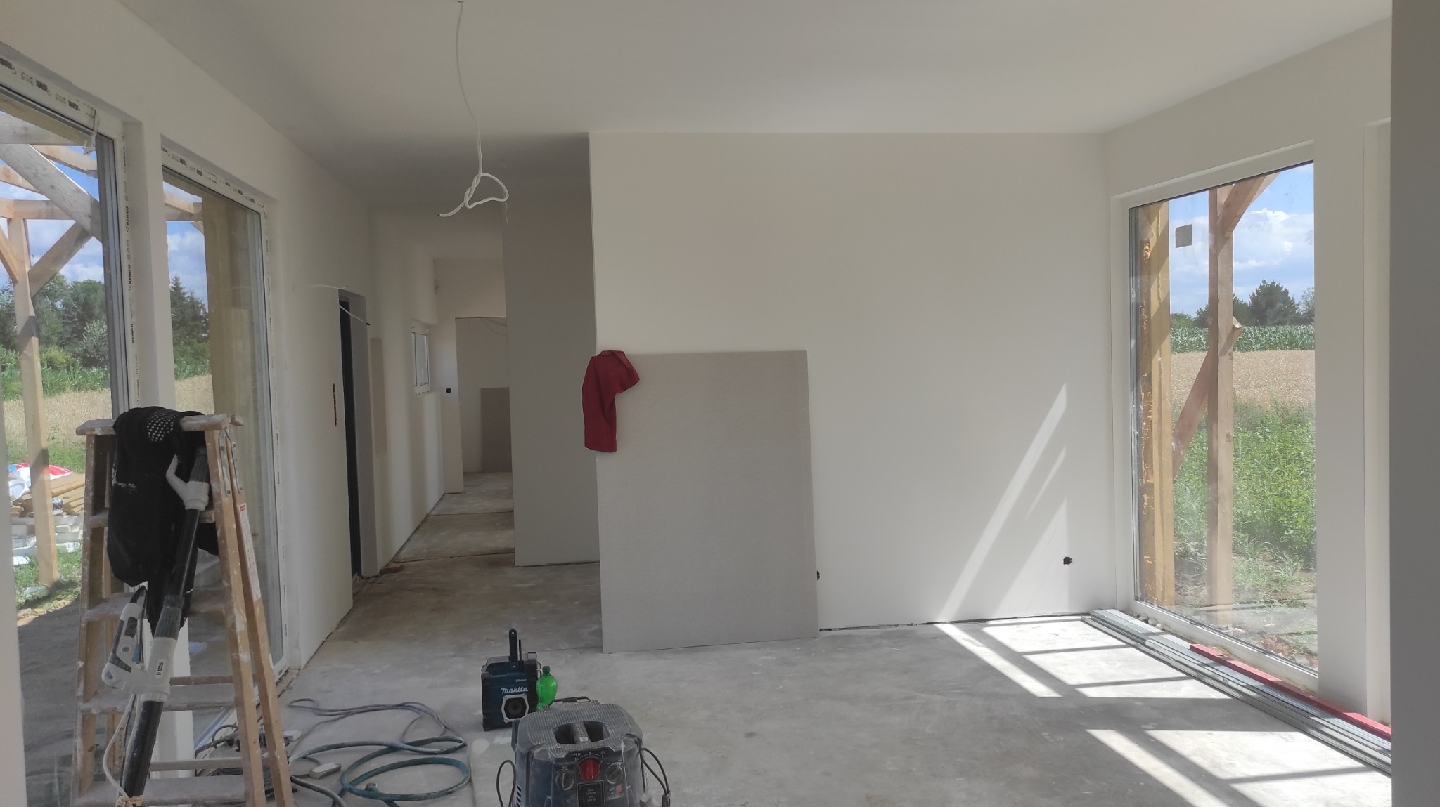 Wnętrze w trakcie prac wykończeniowych: ściany po wstępnym wyrównaniu, widoczne narzędzia (szlifierka, odkurzacz), drabina oparta o okno, płyta karton-gips oparta o ścianę, czerwona szmatka wisząca...