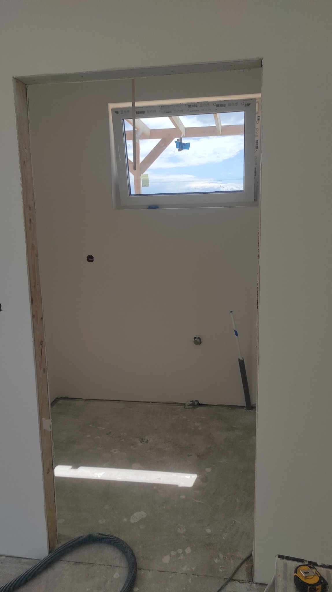 Przygotowane do malowania wnętrze z osadzonym oknem dachowym, widoczne ślady po szpachlowaniu, rurka instalacyjna przy ścianie, oraz wąż od odkurzacza i miarka na podłodze.