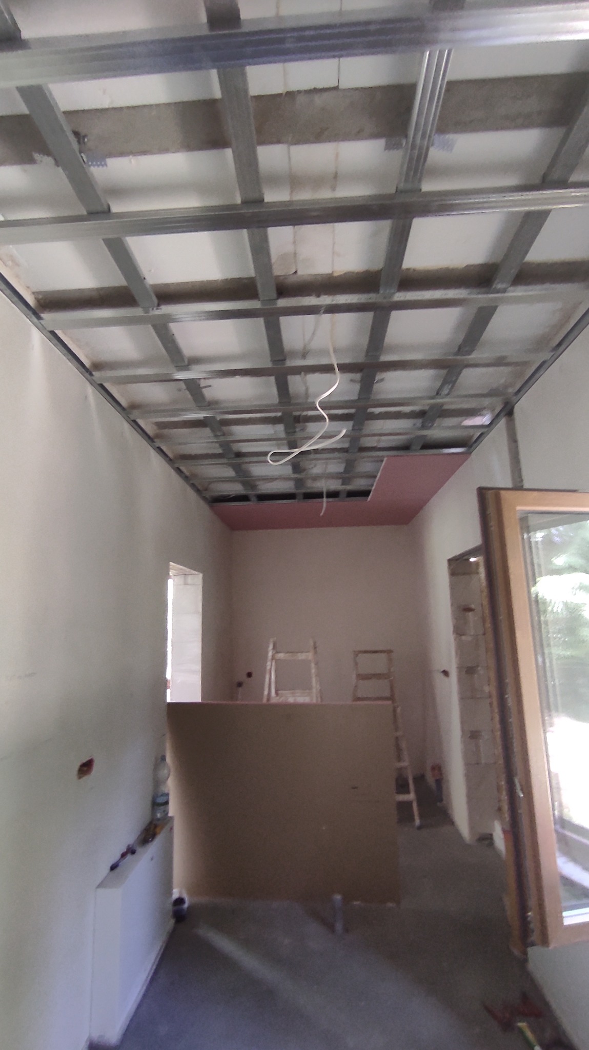 Montaż sufitu podwieszanego: metalowa konstrukcja kratowa zamocowana do stropu, częściowo obłożona płytą karton-gips w kolorze różowym, widoczne kable instalacji elektrycznej, w tle drabiny malarskie.