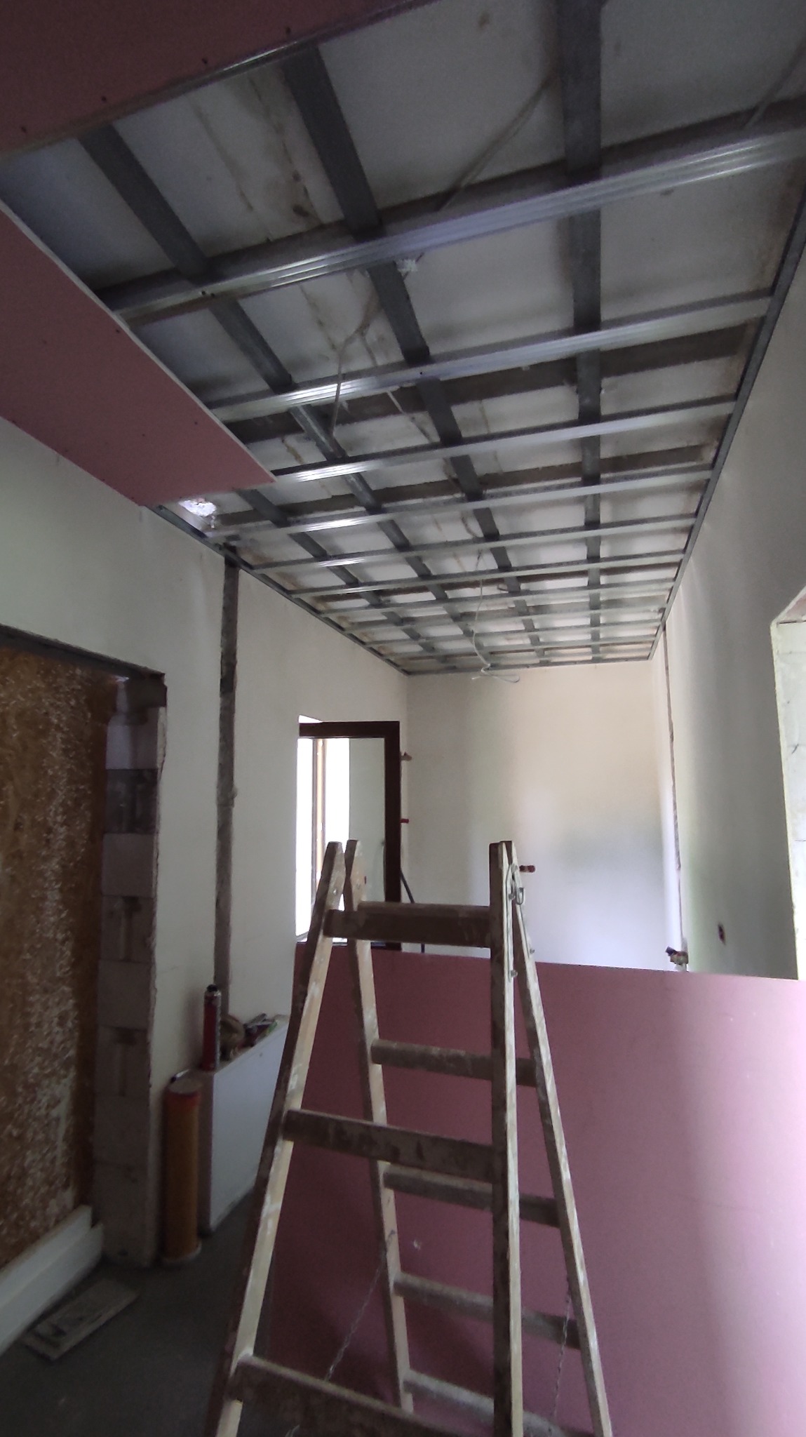 Montaż sufitu podwieszanego z płyt kartonowo-gipsowych w trakcie remontu, widoczna metalowa konstrukcja, częściowo zamontowane płyty oraz drewniana drabina.