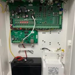 Otwarta obudowa centrali alarmowej Satel Integra Plus z widoczną elektroniką, akumulatorem i transformatorem, przewody uporządkowane w listwach zaciskowych.