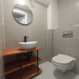 Nowoczesna łazienka z wiszącą toaletą, okrągłym lustrem i umywalką nablatową na drewnianym regale z czarnym kranem, szare płytki na ścianach z subtelnym wzorem.