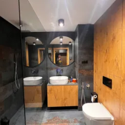 Nowoczesna łazienka z dwoma okrągłymi lustrami, umywalkami nablatowymi na szafkach z drewnopodobnego materiału, toaleta wisząca i kabina prysznicowa z ciemnymi płytkami imitującymi drewno.