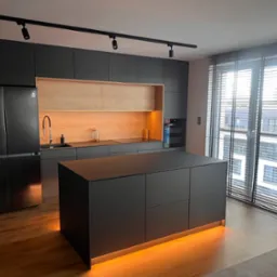 Nowoczesna kuchnia z ciemnymi, matowymi frontami szafek, drewnianym wykończeniem wnęki nad blatem, wyspą kuchenną z podświetleniem LED u dołu, oświetleniem szynowym i drewnianą podłogą.