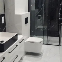 Nowoczesna łazienka w bieli i czerni: marmurowe płytki, podwieszana toaleta, kabina prysznicowa z czarnymi ramami, biała szafka z czarną umywalką.