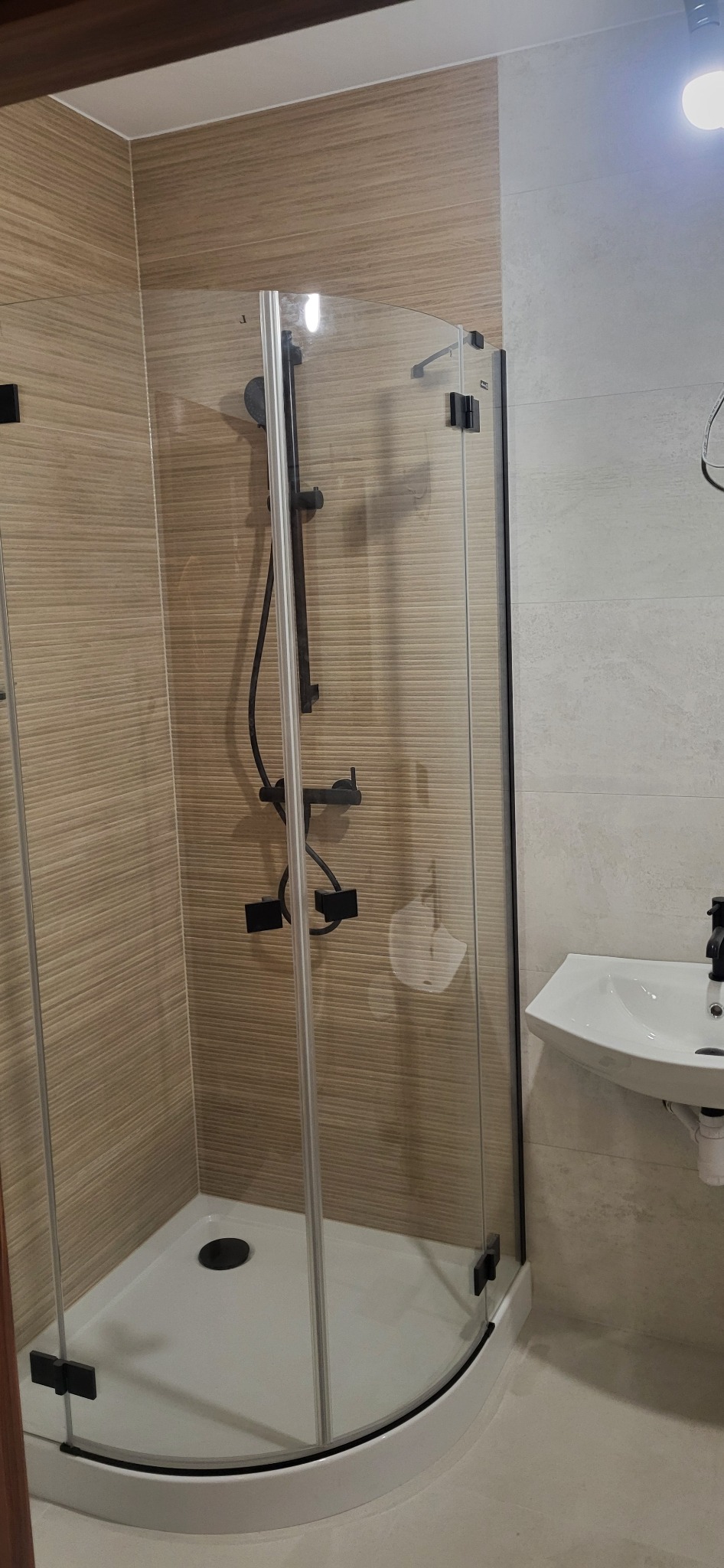 Wykończona kabina prysznicowa z czarną armaturą i odpływem liniowym, umywalka narożna. Beżowe płytki imitujące drewno na ścianach.