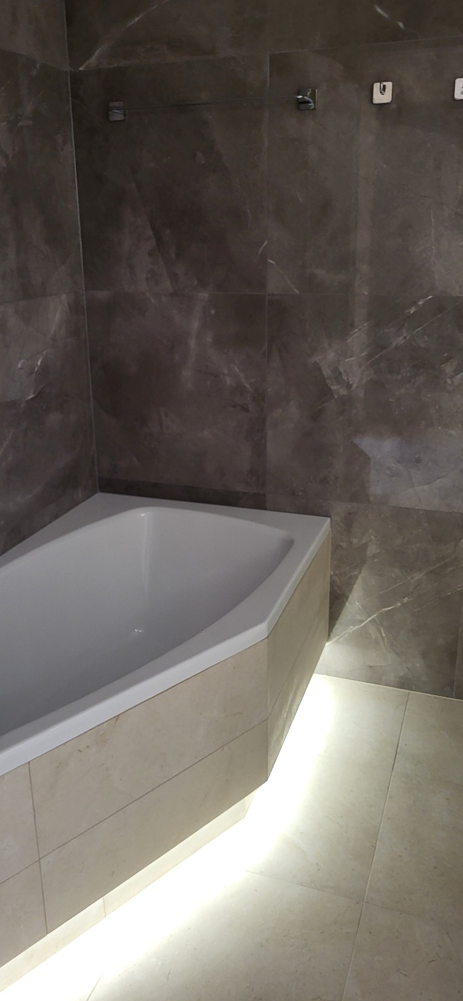 Narożna wanna z podświetleniem LED, obudowana płytkami w odcieniach beżu. Ściany wyłożone marmurowymi płytkami w tonacji szarości. Minimalistyczne wieszaki na ścianie.