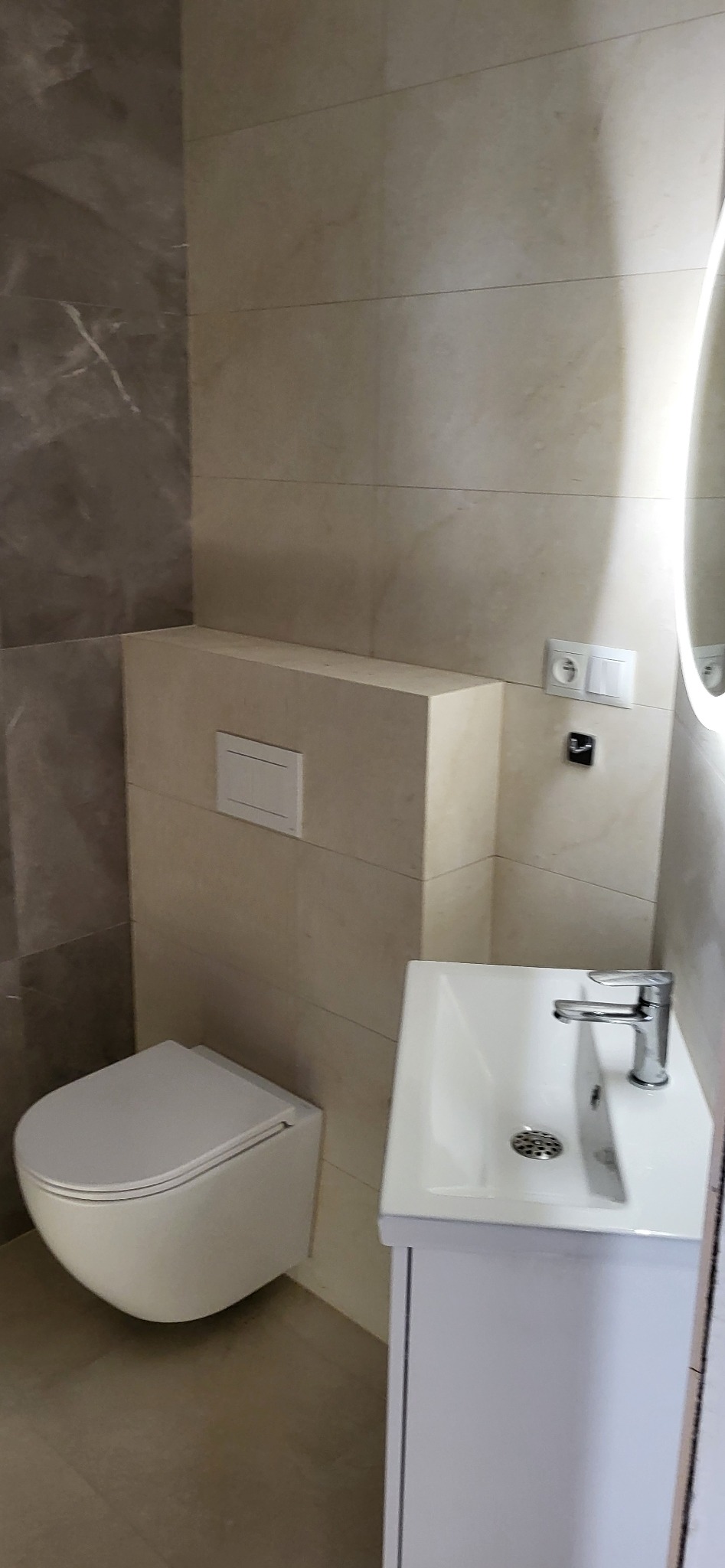 Nowoczesna łazienka w beżach i szarościach: podwieszana toaleta, umywalka z szafką, płytki imitujące marmur, okrągłe lustro z podświetleniem LED i minimalistyczna armatura.