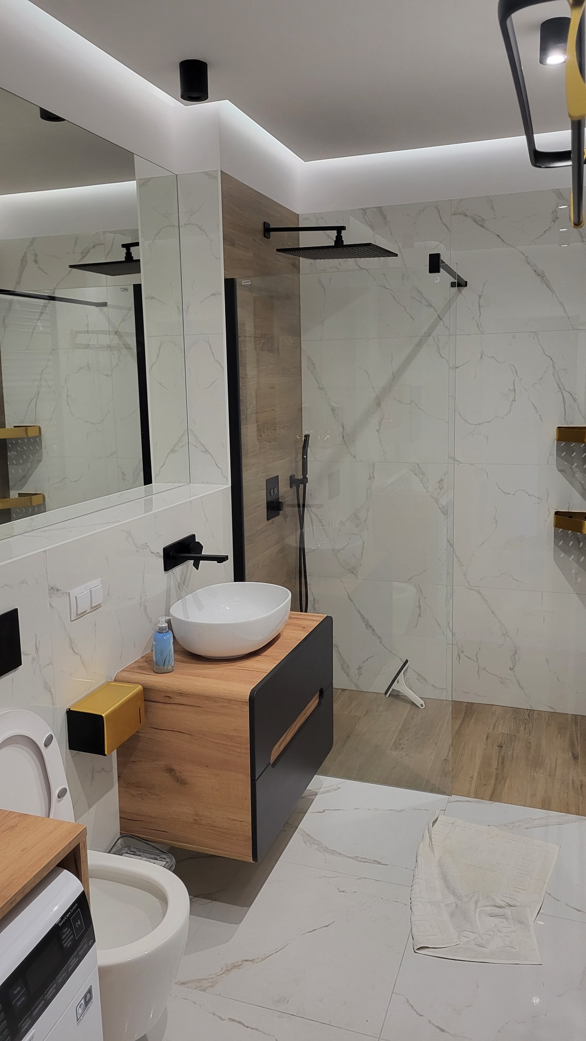 Nowoczesna łazienka z marmurowymi płytkami, drewnianą szafką pod umywalką i czarną armaturą. Kabina prysznicowa typu walk-in z deszczownicą. Oświetlenie LED.