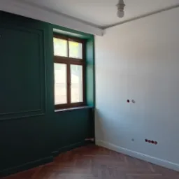 Pokój po odnowieniu: ściana pomalowana na ciemnozielono z dekoracyjnymi panelami, okno w drewnianej ramie, parkiet ułożony w jodełkę, biała ściana z przygotowanymi gniazdkami elektrycznymi, listwy...