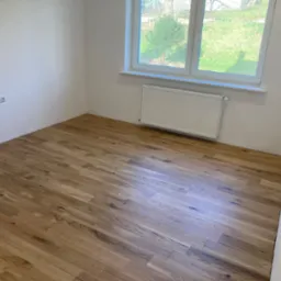 Pusty pokój po remoncie: świeżo położona drewniana podłoga, białe ściany z gniazdkami elektrycznymi, grzejnik pod oknem z widokiem na zieleń.