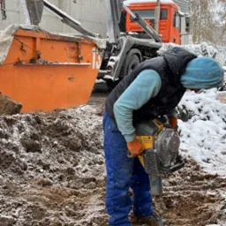 Pracownik w niebieskim ubraniu i pomarańczowych rękawicach używa młota pneumatycznego do rozbijania ziemi, obok stoi pomarańczowa ciężarówka z dźwigiem podczas lekkiego opadu śniegu.