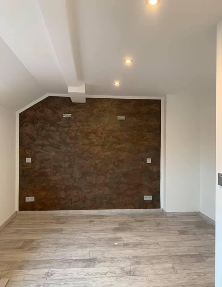 Pokój po remoncie z brązową ścianą o teksturalnym wykończeniu, jasną podłogą imitującą drewno oraz białymi ścianami i sufitem z trzema punktowymi lampami.